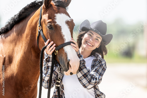 Happy young woman stands next to her horse's head outdoors on a summer day