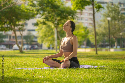 A woman in brown sport bra and black short sitting on yoga mat doing maditation with hand together at chest on grass floor and garden blurry background.