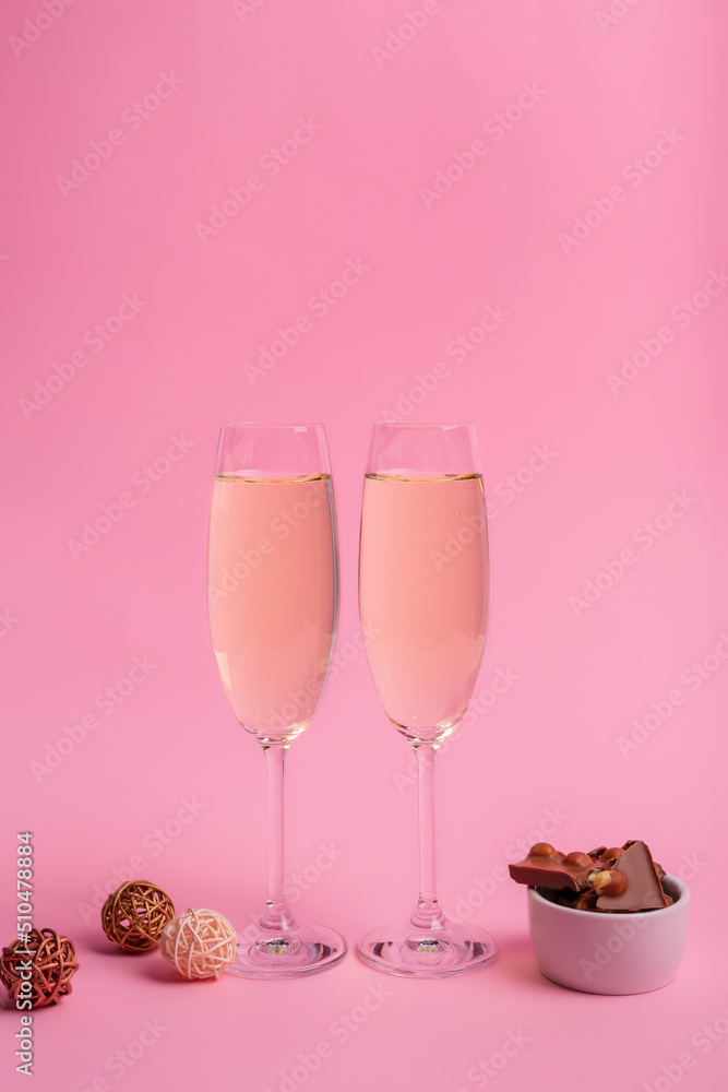 Two glasses of white sparkling wine and tasty milk chocolate with nuts in a small white plate on pink background. Beautiful holiday card. Two glasses of white sparkling wine and tasty milk chocolate with nuts in a small white plate on pink background. Beautiful holiday card.