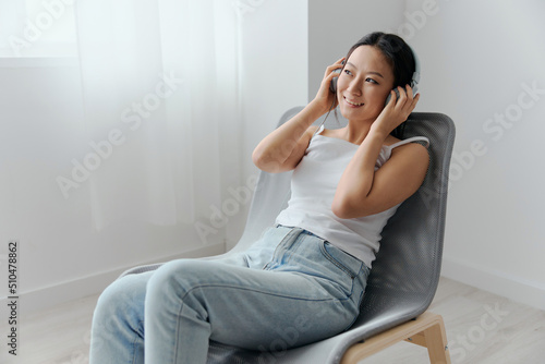 Cheerful dreaming tanned lovely young Asian woman in headphones enjoy favorite playlist looks aside at home interior living room. Sound Studio Stream Social media concept. Cool offer Banner Wide angle