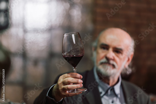 Caucasian senior man in a suit, standing in an old wine cellar with wooden barrels, expertly tasting red wine, checking a color, smell, and taste