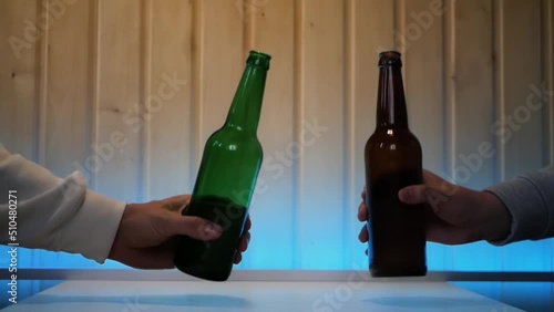 Close up of two male hands clinking a pair of beer bottles in greeting. Two men cheer beers at home in a pleasant atmosphere with blue blacklight. Concept of a toast to friendship with beer drinks photo