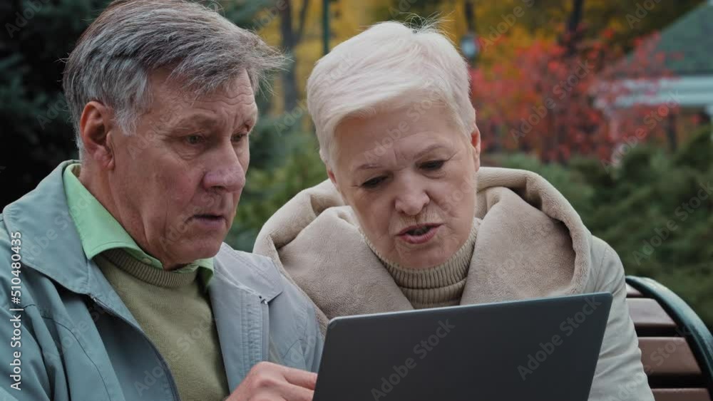 Caucasian senior couple mature wife with husband sit on bench outdoors use wi-fi on laptop elderly spouses look at pc screen shopping online search information news work in web app make internet order Caucasian senior couple mature wife with husband sit on bench outdoors use wi-fi on laptop elderly spouses look at pc screen shopping online search information news work in web app make internet order