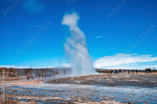 Tourists enjoying Strokkur geyser eruption. Smoke emitting from dramatic landscape against blue sky. Men and women exploring famous attraction in valley during sunny day.
