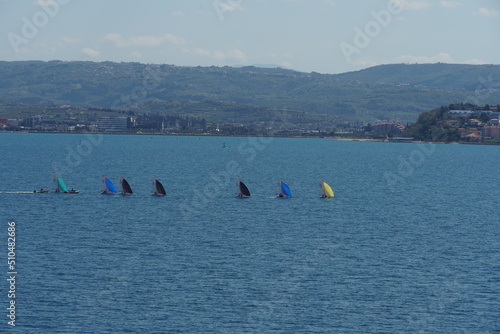 Small sailing boats with different colors sails passing through bay in front of port of Koper in Slovenia during calm weather. In the horizon is mountain coast of Slovenia and buildings of the town.