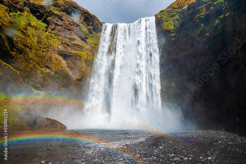 Low angle view of beautiful rainbows against powerful cascades of Skogafoss waterfall. River flowing from mountain in volcanic valley. Scenic natural wilderness of landscape against cloudy sky.
