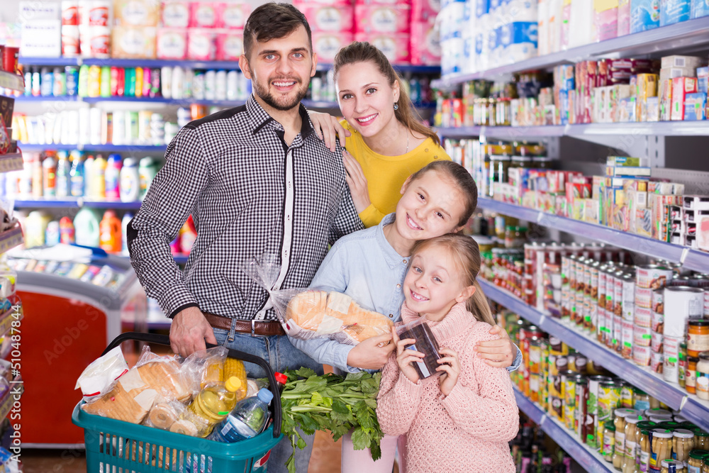Cheerful family of buyers holding full basket after shopping in food store
