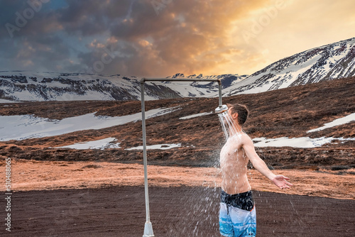 Young tourist showering in hot shower station from geothermal power at Krafla. Shirtless traveler enjoying bath against snow covered mountain. He is spending leisure time against sky during sunset. photo