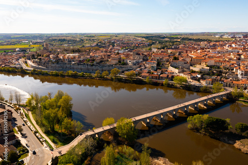 Picturesque aerial view of Zamora city overlooking brownish tiled roofs of residential buildings and ancient arched bridge over Duero river on spring day, Spain