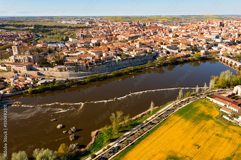 Picturesque aerial view of Zamora city overlooking brownish tiled roofs of residential buildings and ancient arched bridge over Duero river on spring day, Spain