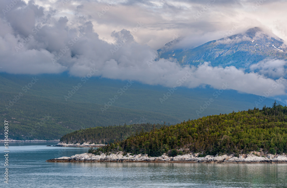 Skagway, Alaska, USA - July 20, 2011: Taiya Inlet above Chilkoot Inlet. Gray cloudscape descends over forested mountain flanks. White-gray rocky shoreline over blueish water landscape.
