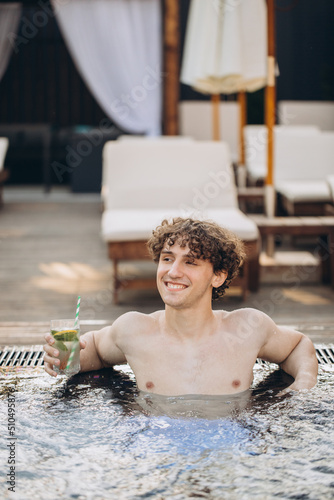 Curly positive guy holds cocktails and enjoys spending time in the jacuzzi