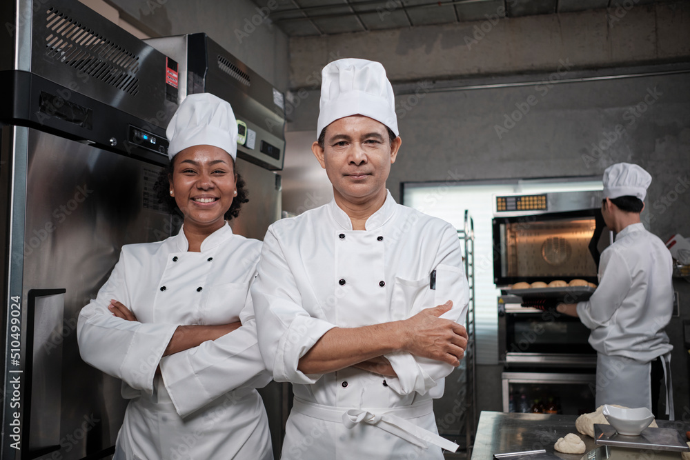 Pastry foods professional partners, two happy chefs team staff in white cooking uniforms stand, arms crossed with confidence, cheerful smiles with commercial culinary jobs in restaurant kitchen.