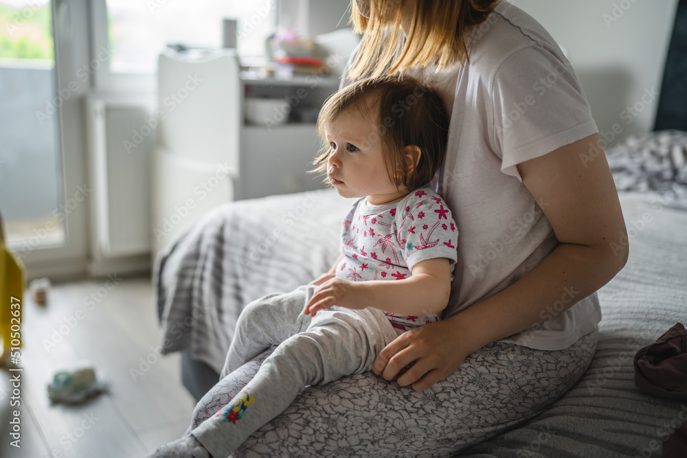 One small caucasian girl toddler sitting on the bed at home while her mother is changing her clothes real people growing up copy space One small caucasian girl toddler sitting on the bed at home while her mother is changing her clothes real people growing up copy space