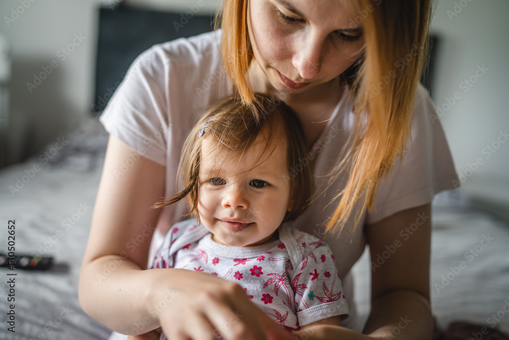 One small caucasian girl toddler sitting on the bed at home while her mother is changing her clothes real people growing up copy space One small caucasian girl toddler sitting on the bed at home while her mother is changing her clothes real people growing up copy space