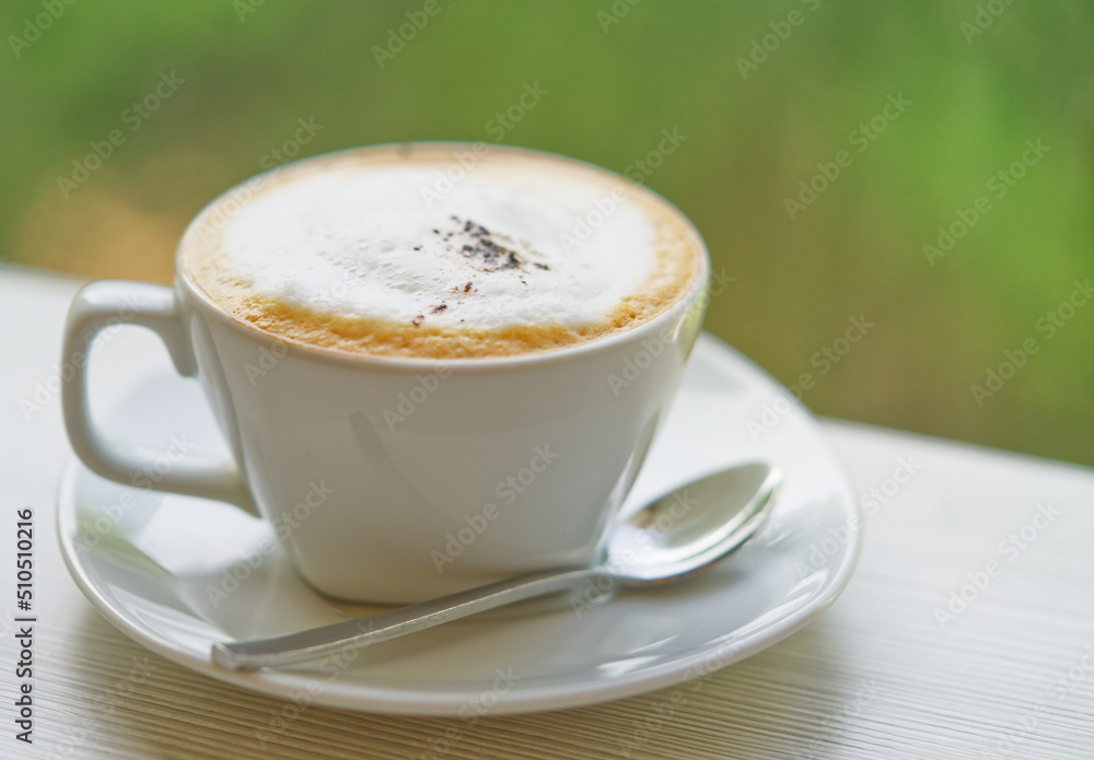 Obraz premium Close up hot cappuccino coffee with natural light, blurred background of green color of tree, selected focus. Small spoon is beside the cup.
