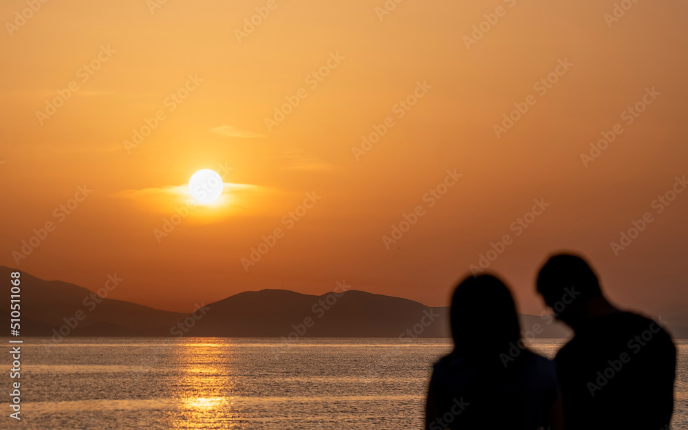 A couple at a sea beach with colorful sunset sky. Tropical beach and seascape and a distant island in the background. Orange and golden sunset sky, relaxing sunlight, summer mood.