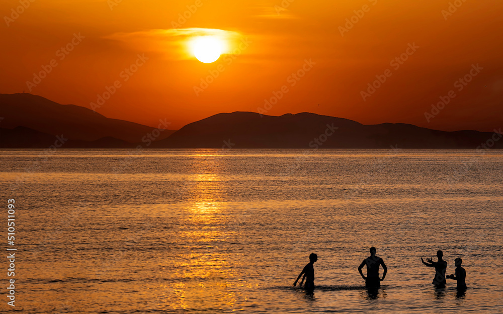 Four friends near the sea beach with colorful sunset sky. Tropical beach and seascape and a distant island in the background. Orange and golden sunset sky, tranquil, relaxing sunlight, summer mood.