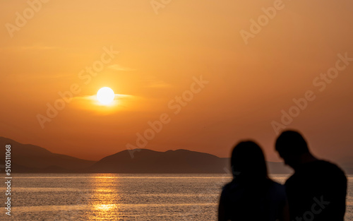 A couple at a sea beach with colorful sunset sky. Tropical beach and seascape and a distant island in the background. Orange and golden sunset sky, relaxing sunlight, summer mood. © Dimitrios