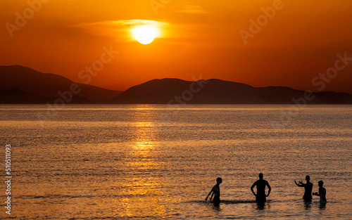 Four friends near the sea beach with colorful sunset sky. Tropical beach and seascape and a distant island in the background. Orange and golden sunset sky, tranquil, relaxing sunlight, summer mood.