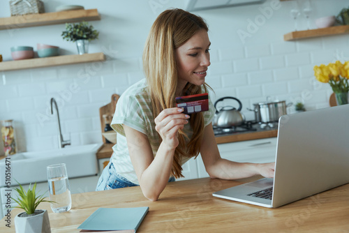 Beautiful young woman enjoying online shopping and using credit card while sitting at the kitchen