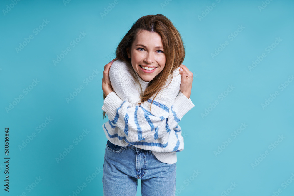 Beautiful young woman hugging self and smiling while standing against blue background Beautiful young woman hugging self and smiling while standing against blue background