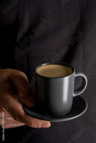 A man's hand holds a cup of hot espresso coffee against the background of a black T-shirt.