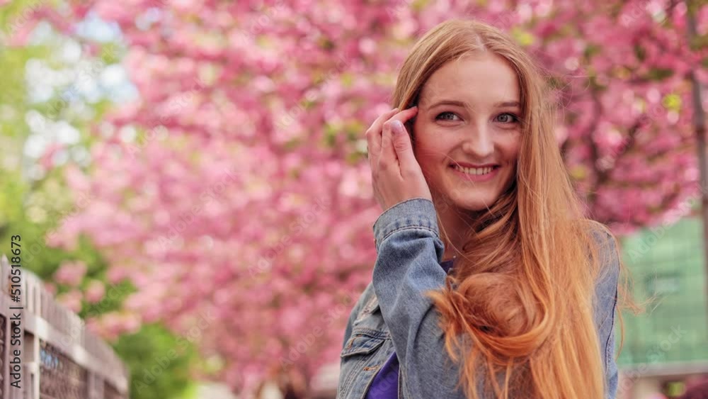 Happy Caucasian female with long hair in casual denim jacket walking around Cherry blossom trees. Portrait of young woman look around and smiling on blurred background in the city street. Sakura tree. Happy Caucasian female with long hair in casual denim jacket walking around Cherry blossom trees. Portrait of young woman look around and smiling on blurred background in the city street. Sakura tree.