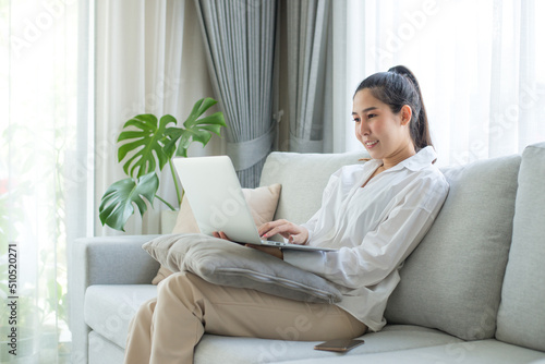 Image of cheerful asian woman using laptop while sitting on couch in living room. She uses her laptop for meetings  searching for information  chatting with friends  and enjoying shopping online.
