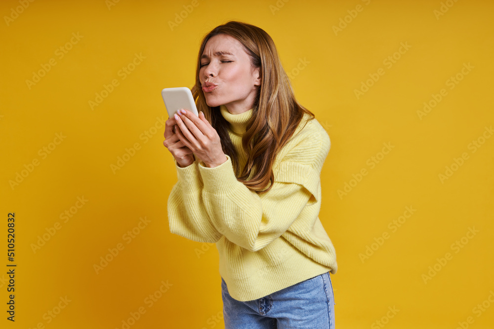 Beautiful woman holding smart phone and sending a kiss while standing against yellow background Beautiful woman holding smart phone and sending a kiss while standing against yellow background