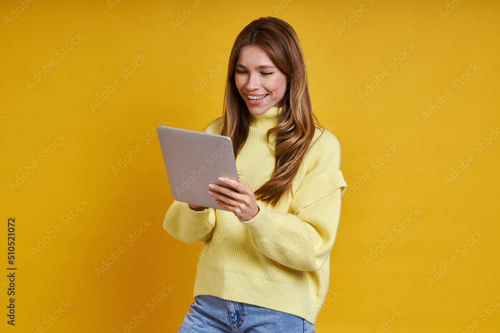 Happy young woman working on digital tablet while standing against yellow background Happy young woman working on digital tablet while standing against yellow background