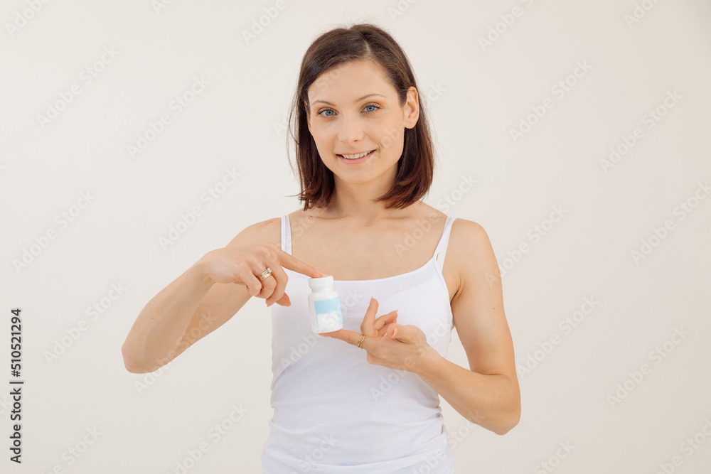 Young smiling woman advertising useful vitamins for women closeup, white background. Holding in hands and pointing at jar of pills, disease treatment. Female health and reproductive capacity concept Young smiling woman advertising useful vitamins for women closeup, white background. Holding in hands and pointing at jar of pills, disease treatment. Female health and reproductive capacity concept