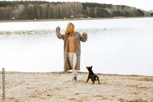 Cheerful young blonde woman in warm clothes playing and having fun with cute puppy on sandy river bank on cool sunny day. Outdoors weekend activity, dogginess, water scenery background photo