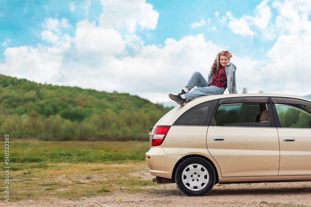 Pretty teen girl is sitting on the roof of a car. Mountains and sky are in the background. Copy space. The concept of freedom and obtaining a driver's license