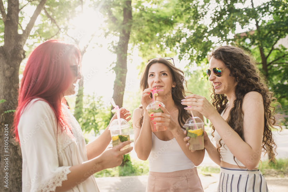 Three young women in fashionable clothes and bright sunglasses walk around the city in summer and drink refreshing non-alcoholic cocktails from disposable plastic cups. Women's friendship and walks in Three young women in fashionable clothes and bright sunglasses walk around the city in summer and drink refreshing non-alcoholic cocktails from disposable plastic cups. Women's friendship and walks in