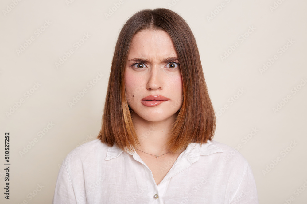 Portrait of dark haired Caucasian attractive woman wearing shirt posing isolated over white background, looking at camera with frowning face, being confused.