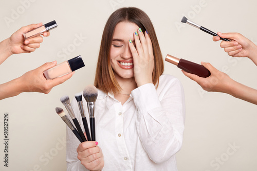 Photos Image of the hands of several beauticians holding their respective equipment giving makeover to beautiful smiling woman with charming smile, standing and covering eye with palm