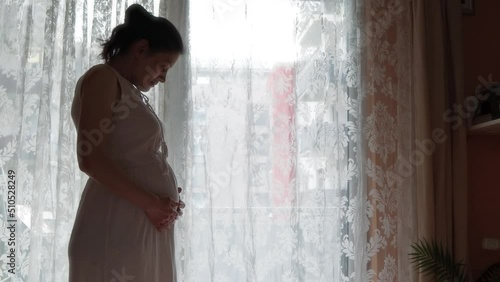 Pregnant woman by the window thinking and playing with baby clothes