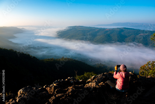 Young traveler woman with mobile phone sitting on the rock to take a photo of sunrise landscape scenery in the morning. 
