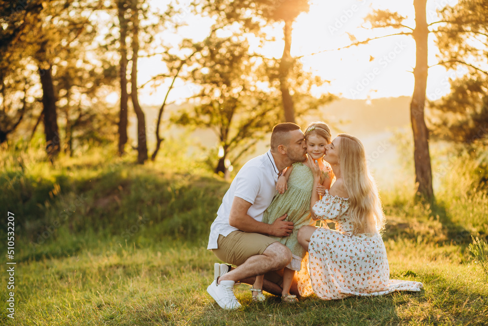 Fototapeta premium Happy family in the park evening light. The lights of a sun. Mom, dad and baby happy walk at sunset. The concept of a happy family.Parents hold the baby's hands.