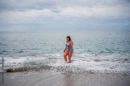 A plump woman in a bathing suit enters the water during the surf. Alone on the beach, Gray sky in the clouds, swimming in winter.