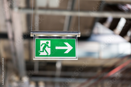 Emergency exit sign. With running figure and arrow to the right. Blurry background, hanging from the ceiling.