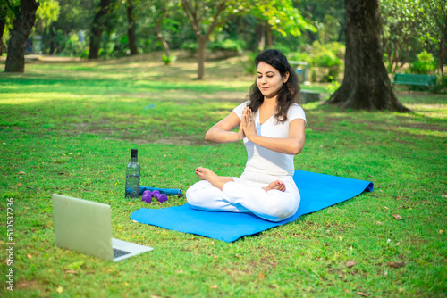 Beautiful indian woman doing breathing yoga exercise in the park while using laptop for online class or virtual tutorials, Asian female meditation pose, healthcare. online learning