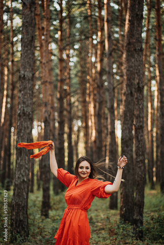Barefoot happy young woman in red dress with hand raised dancing in pine forest at summer day.