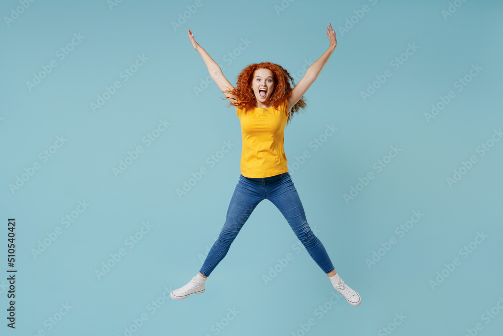 Full body young excited happy redhead woman 20s wearing yellow t-shirt jump high with outstretched legs hands isolated on plain light pastel blue background studio portrait. People lifestyle concept.