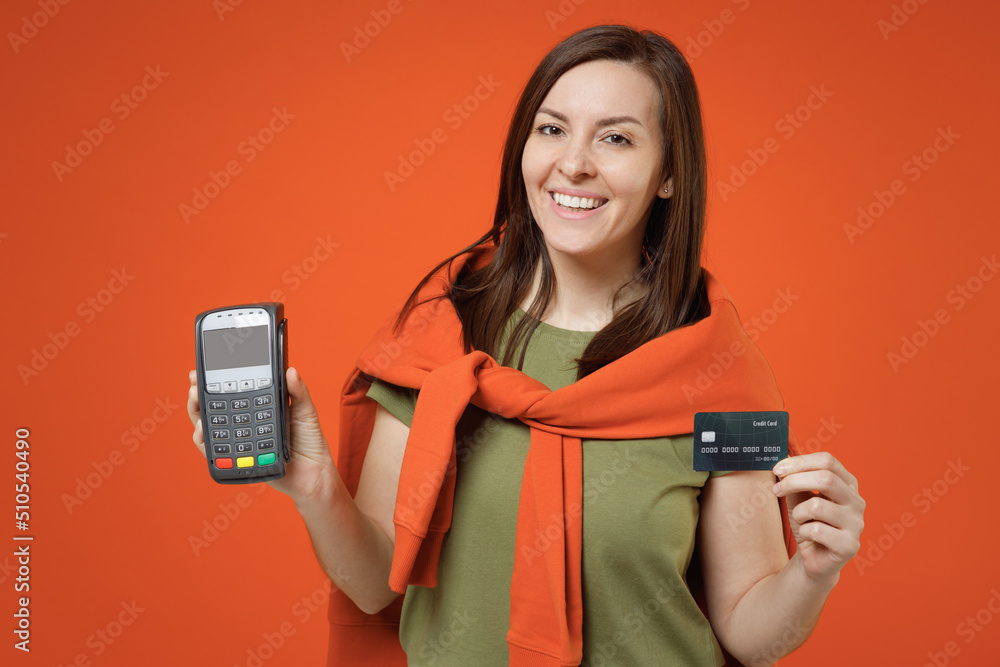 Young smiling happy woman 20s in khaki t-shirt tied sweater on shoulders hold wireless modern bank payment terminal to process acquire credit card payments isolated on plain orange background studio. Young smiling happy woman 20s in khaki t-shirt tied sweater on shoulders hold wireless modern bank payment terminal to process acquire credit card payments isolated on plain orange background studio.