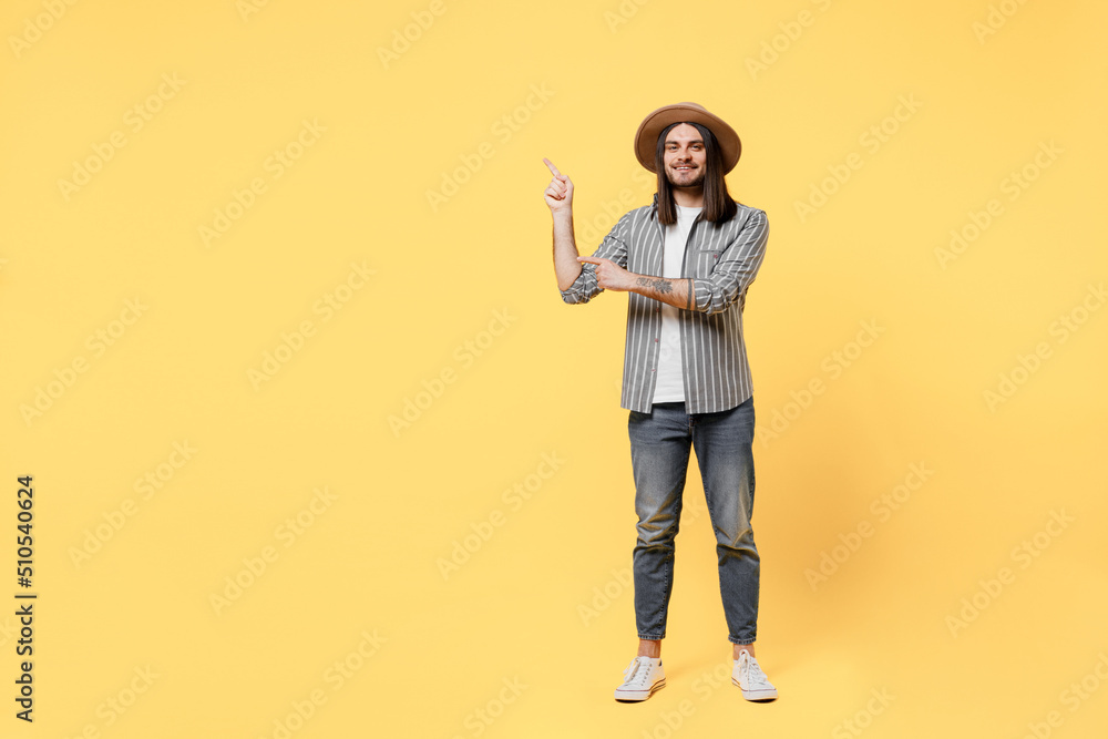 Fototapeta premium Full body happy young man he 20s in striped grey shirt white t-shirt hat pointing index finger aside indicate on workspace area copy space mock up isolated on plain yellow background studio portrait
