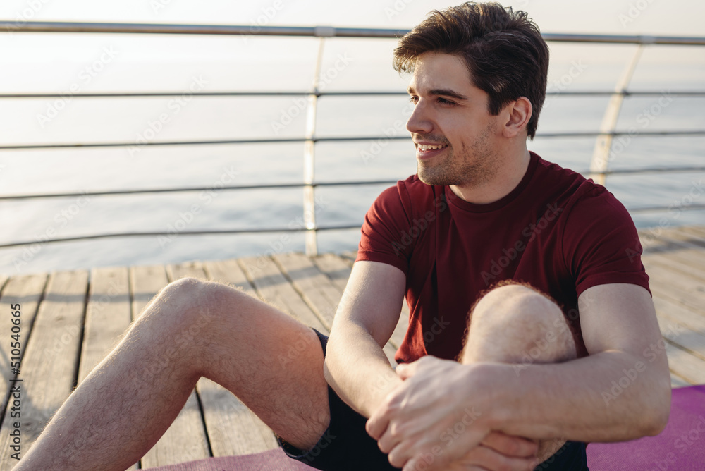 Young strong sporty athletic toned fit sportsman man wear sports clothes sit on yoga mat look aside warm up training at sunrise sun dawn over sea beach outdoor on pier seaside in summer day morning. Young strong sporty athletic toned fit sportsman man wear sports clothes sit on yoga mat look aside warm up training at sunrise sun dawn over sea beach outdoor on pier seaside in summer day morning.