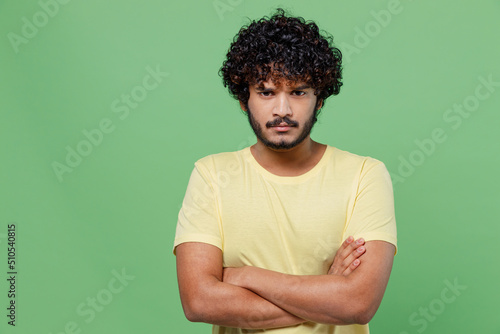 Young unhappy offended indignant sad Indian man 20s in yellow t-shirt hold hands crossed folded look camera isolated on plain pastel light green background studio portrait. People lifestyle concept.