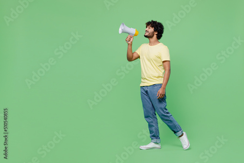 Full size young smiling happy Indian man 20s in basic yellow t-shirt hold scream in megaphone announces discounts sale Hurry up isolated on plain pastel light green background People lifestyle concept